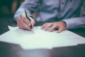 A person in a long-sleeved dress shirt writes with a pen on white paper.