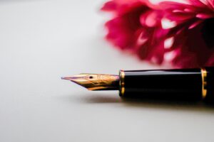 A black fountain pen with a gold nib rests on a grey surface with a red flower blurred in the background. 